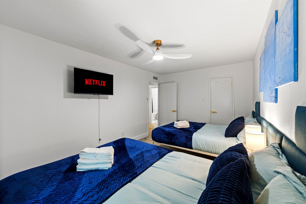 Spacious bedroom with two plush queen beds, blackout curtains, and a smart TV—ideal for families or groups to unwind in comfort.