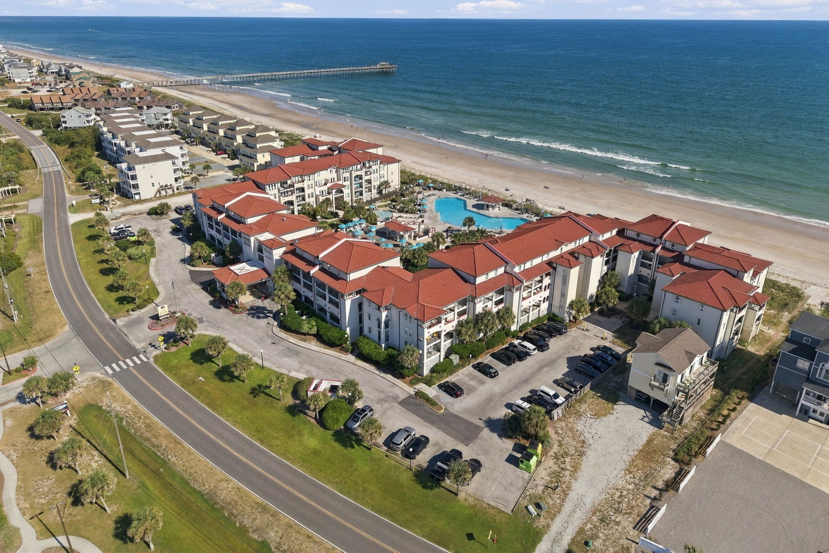 Aerial view of the oceanfront resort grounds, highlighted by terracotta-roofed buildings, beach access, parking areas, and an adjacent pier stretching into the ocean.   