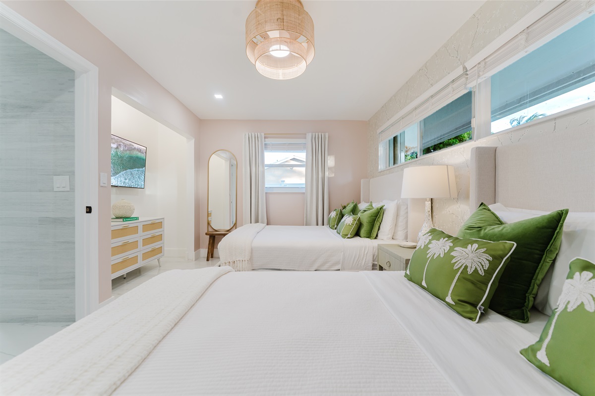Bright, serene guest suite with two queen beds, hotel-quality linens, a wall-mounted smart TV, and ample storage. Features blackout curtains and blinds, a rattan ceiling fan, and a private en-suite bathroom—ideal for families or guests sharing space.