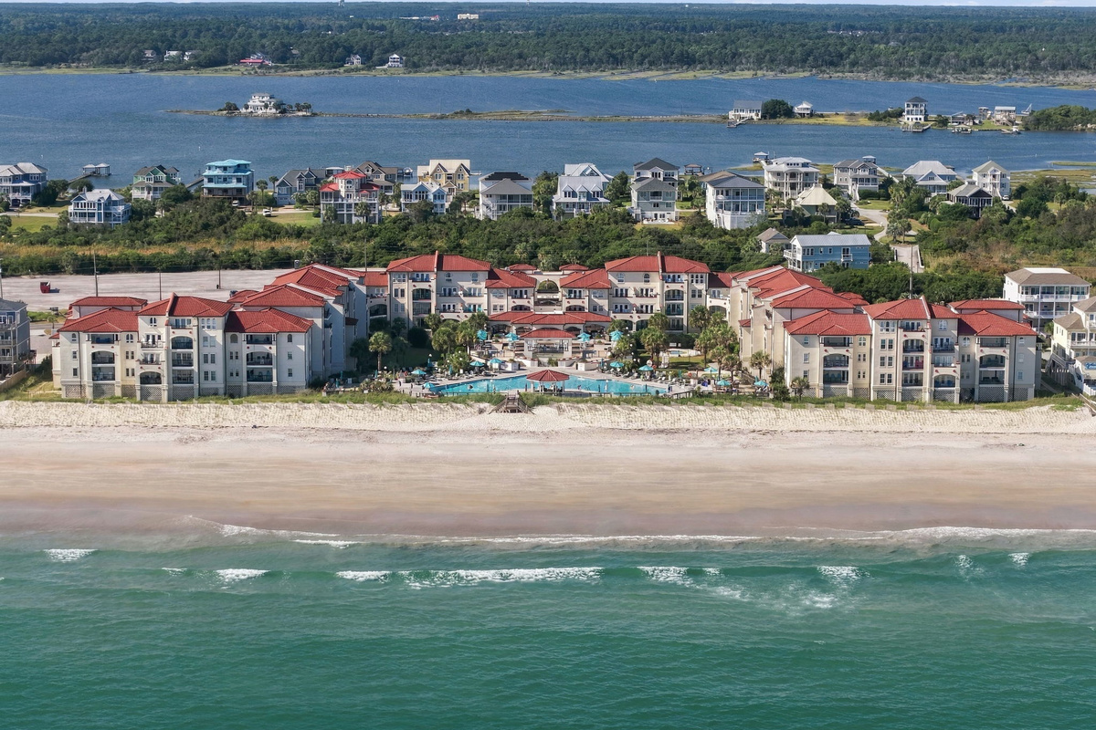 Direct oceanfront aerial view of the resort complex, framed by ocean waves on one side and sound waters on the other.   