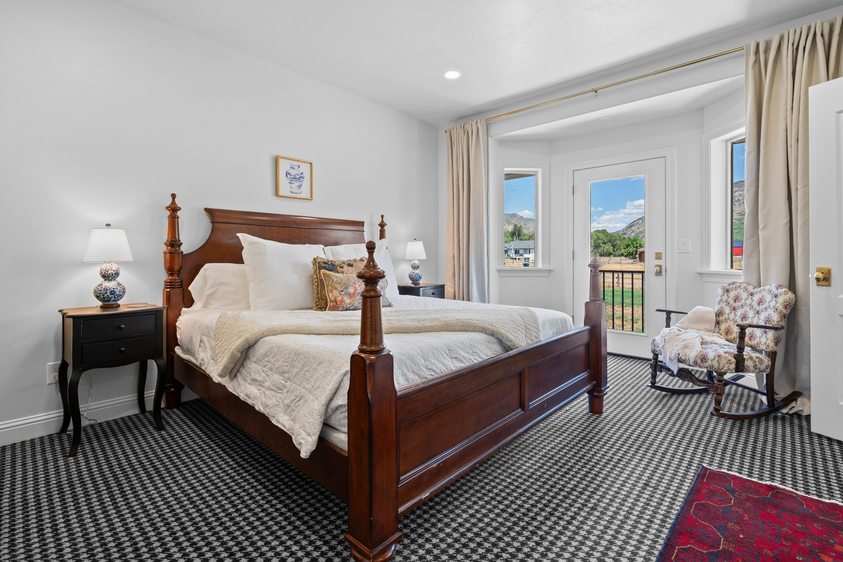 The spacious primary bedroom features a wooden bed frame, layered luxury bedding, a charming sitting area, and private access to a balcony overlooking the surrounding scenery.