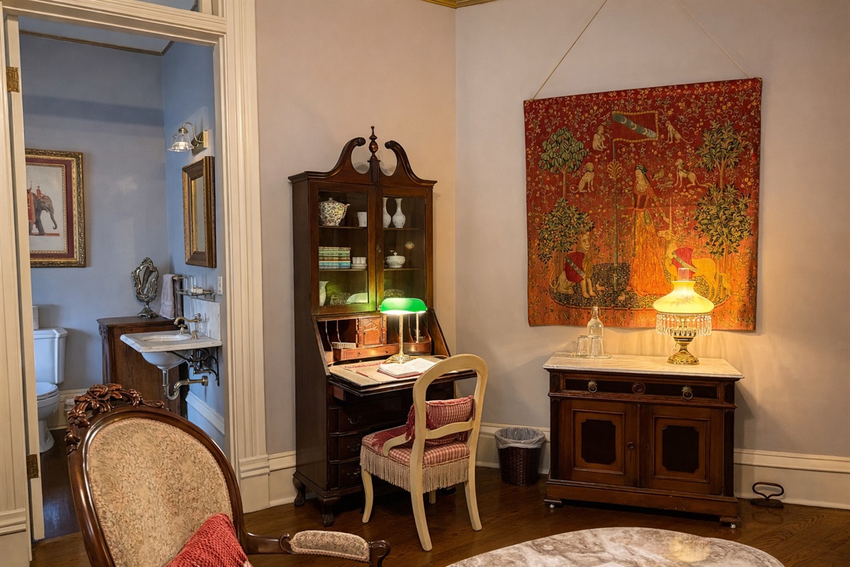 Writing desk area with antique secretary, chair, table lamp, and decorative wall tapestry.