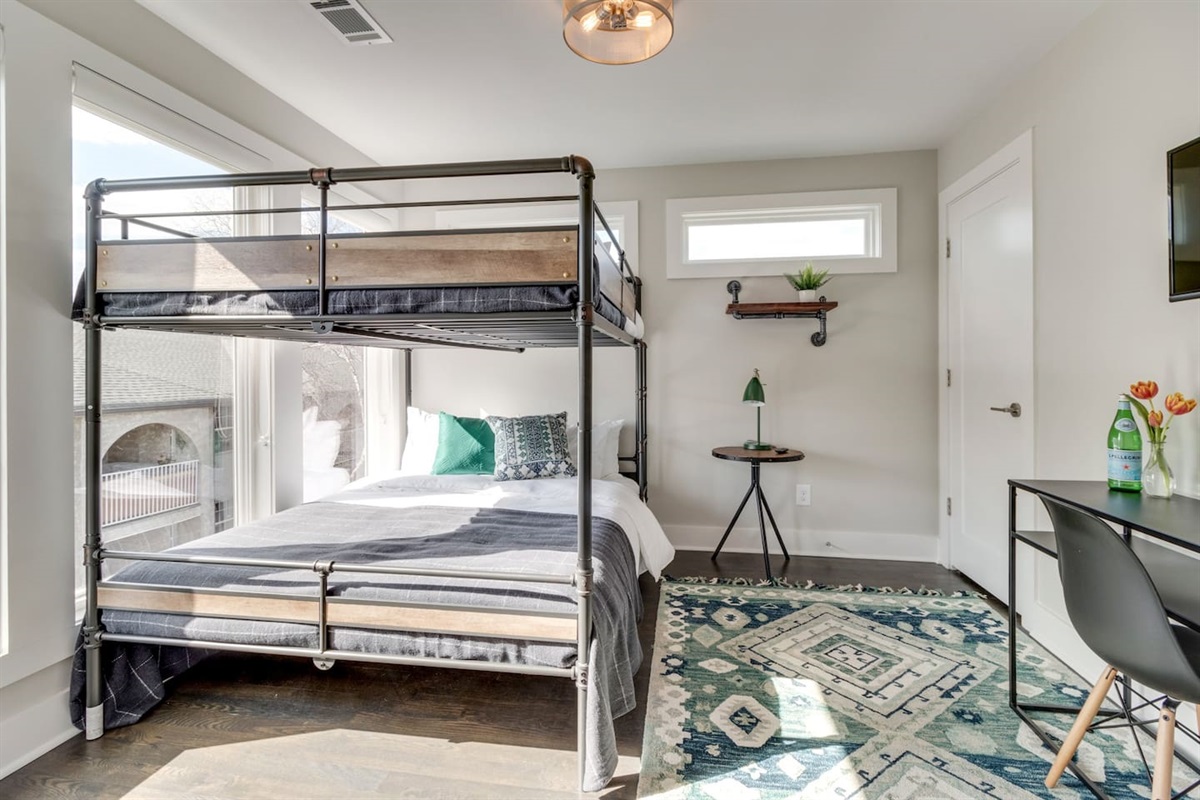 - Industrial style bunk with full bed on bottom and twin up top, styled in cozy neutrals - Bright room with oversized windows, geometric rug, and desk setup for work or study - Thoughtful decor with greenery, light wood accents, and clean minimalist lines