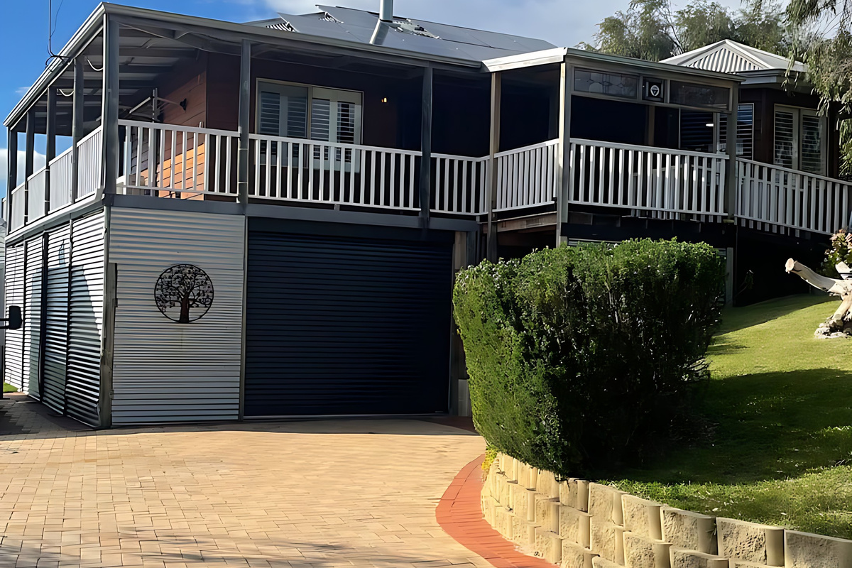 A brick-paved driveway leads up to the garage beneath the house, with the white-railed veranda above and a sloping front lawn, on the edge of Yalgorup National Park at Preston Beach.