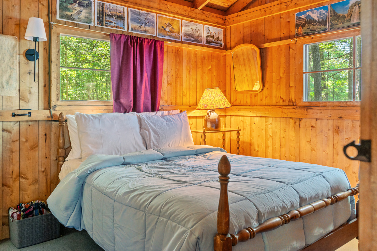 Primary bedroom at Loonrise Point! Features a cozy bed with wooden spindle frame, bedside reading lamps, dual aspect windows overlooking forest greenery, & classic pine walling. Tranquil retreat on Beech Hill Pond in Otis, ME.