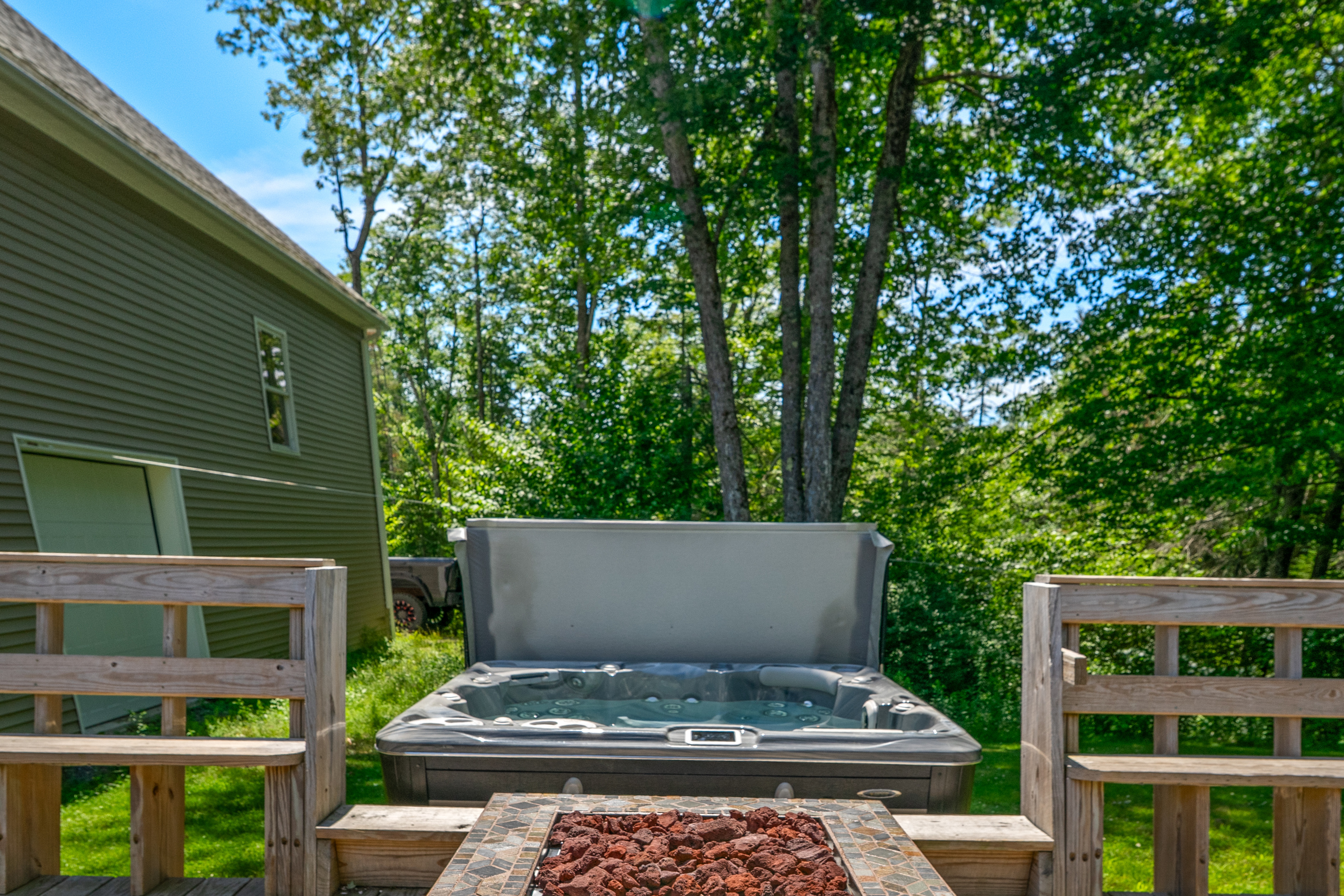 Sit by the propane fire table and look out toward the private 6-person hot tub. Framed by integrated wooden bench seating, this outdoor entertainment setup offers an open view of the quiet, wooded backyard space right off the deck.