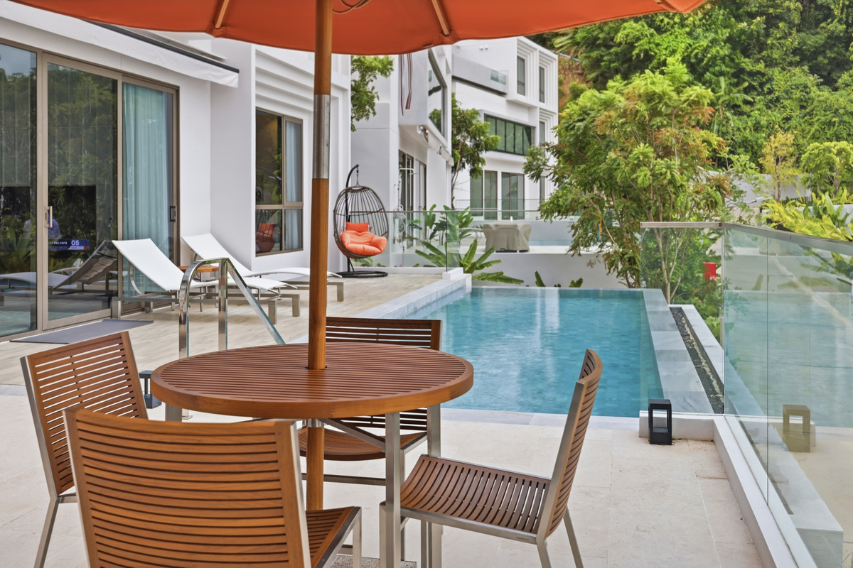 Patio with wooden dining table, lounge chairs, orange umbrella, adjacent pool, and white modern building with greenery.