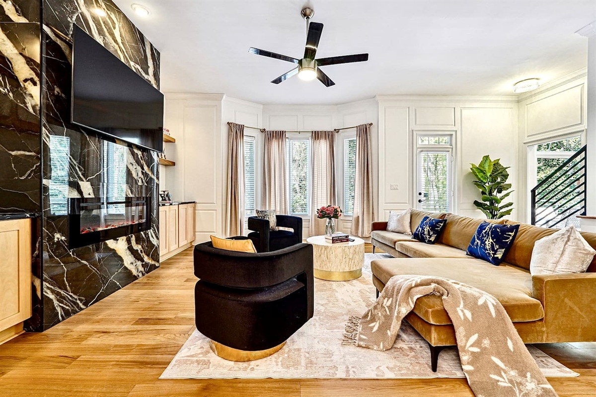 Situated next to the kitchen, this main-floor living room offers a warm, upscale ambiance with a brown L-shaped sectional, two black accent chairs, gold and black design accents, a smart TV, and a fireplace, perfect for entertaining or unwinding.