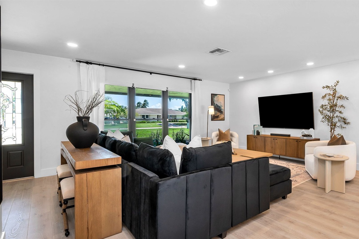 Elegant open-concept living room featuring a plush black velvet sectional, 75” smart TV, and designer mid-century modern furnishings. Floor-to-ceiling windows and French doors fill the space with natural light and connect seamlessly to the outdoor pool.