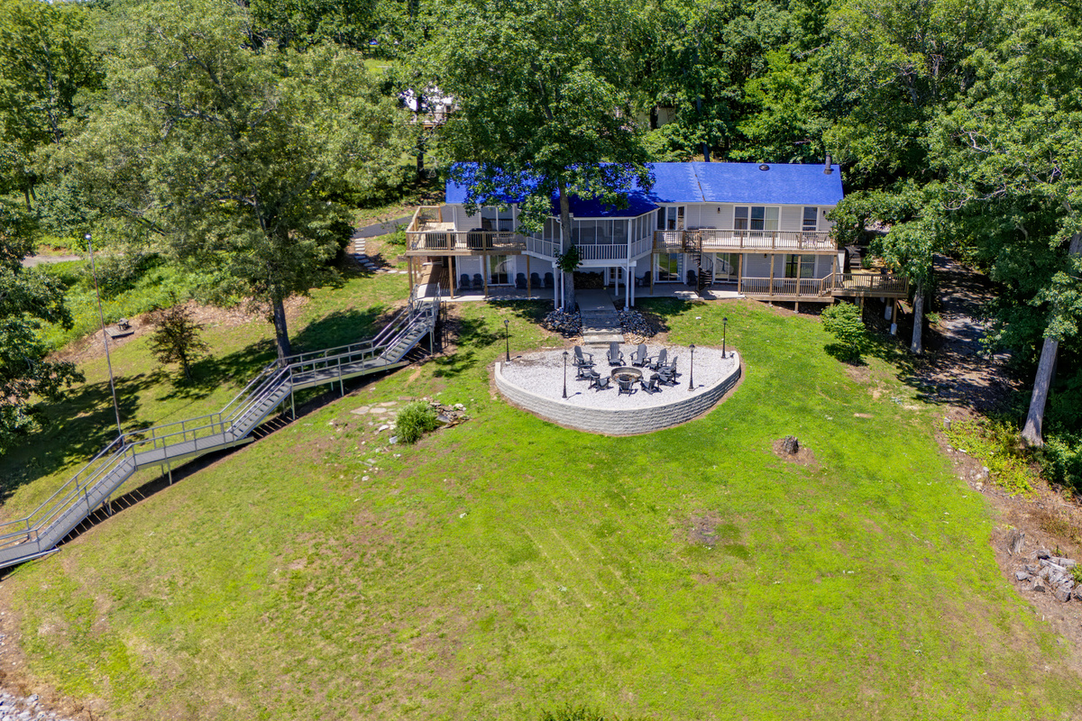A close aerial highlights the home, spacious lawn, circular firepit area, and walkway toward the private dock.