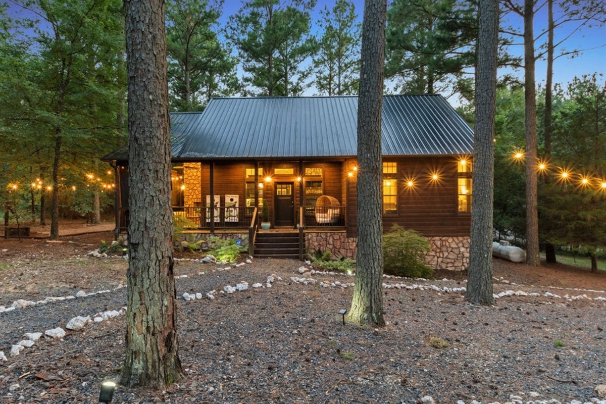 The cabin exterior transforms at dusk with sparkling lights and forest views, creating a cozy, romantic retreat.