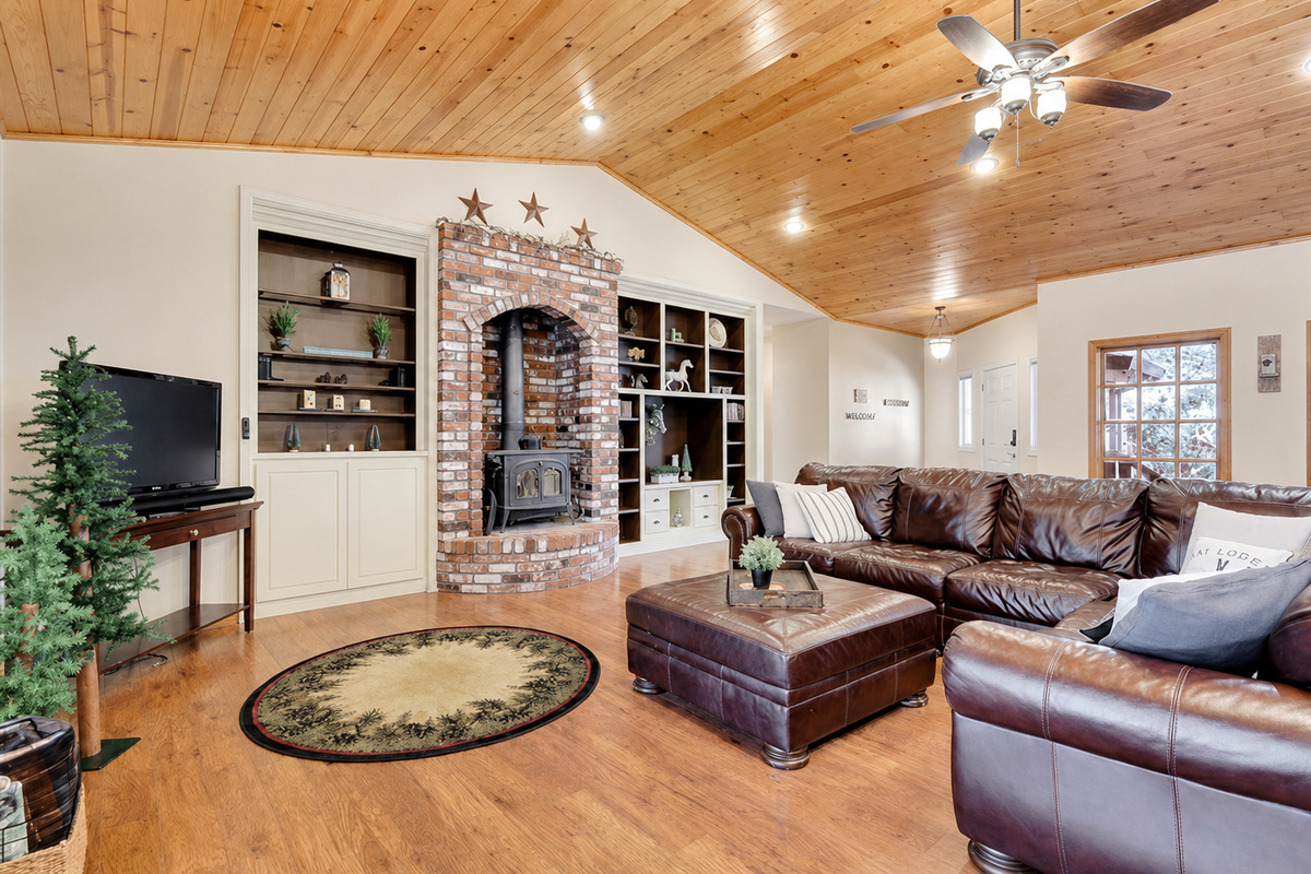 Living room with vaulted wood ceiling, leather sectional seating, brick fireplace, built-in shelving, and TV for cozy cabin downtime.