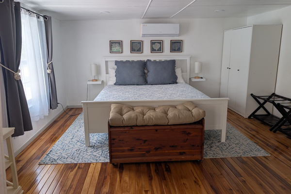 Upstairs master bedroom suite at Villa Coral featuring a white wooden king bed frame, blue coastal linens, rich hardwood flooring, a cedar chest bench, and a split-unit air conditioner in Dorado, Puerto Rico.
