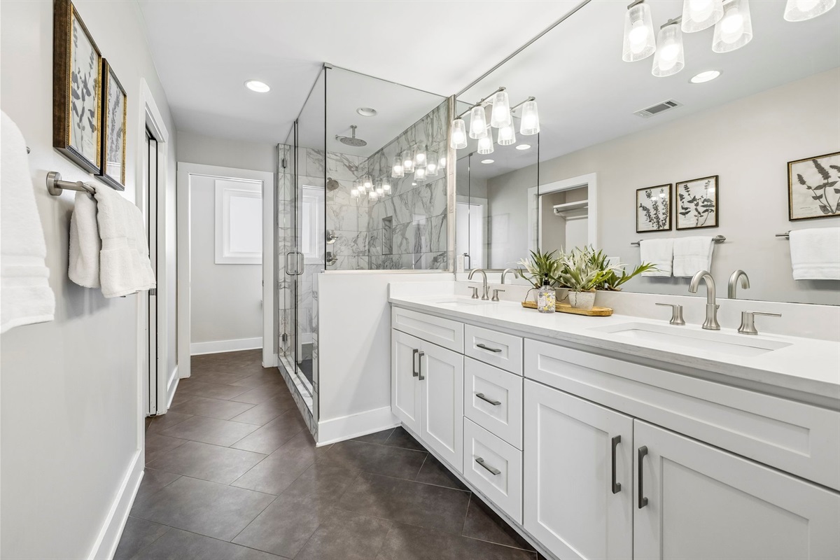 Double sink vanity with modern brushed nickel fixtures and full wall mirror design - Elegant lighting and decorative greenery bring warmth to the sleek, clean space - Glass enclosed shower and easy layout offer a spa like experience with everyday ease