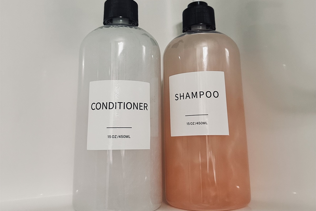 A clear bottle labeled "CONDITIONER" is positioned next to a pink bottle labeled "SHAMPOO," both containing 15 oz (450 ml) of liquid, against a light-colored background.