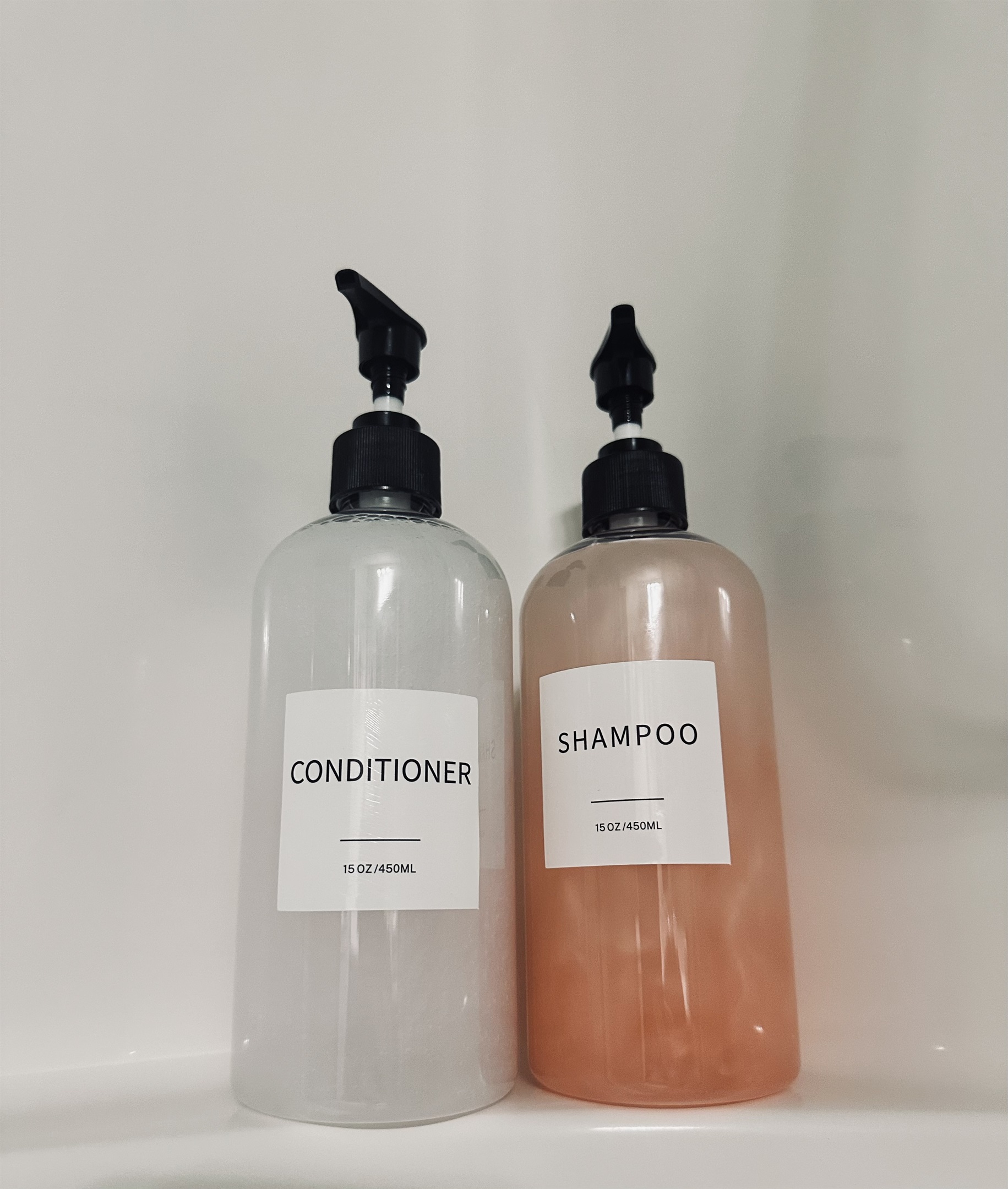 Two transparent bottles with black pump dispensers, labeled "CONDITIONER" and "SHAMPOO," are positioned side by side on a white shelf, each containing a different color liquid—clear for the conditioner and light pink for the shampoo.