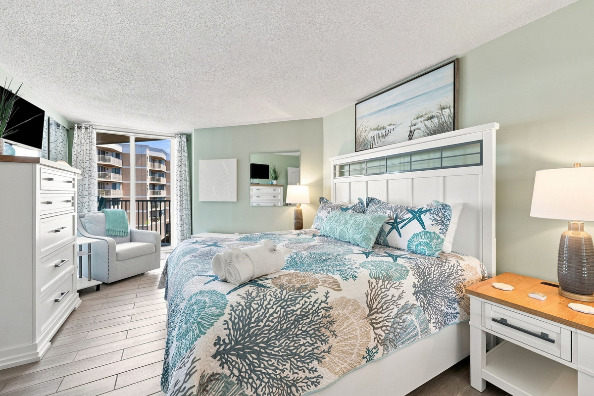 Primary bedroom suite with a cozy king-size bed, coastal bedding, and a large window offering glimpses of the ocean.