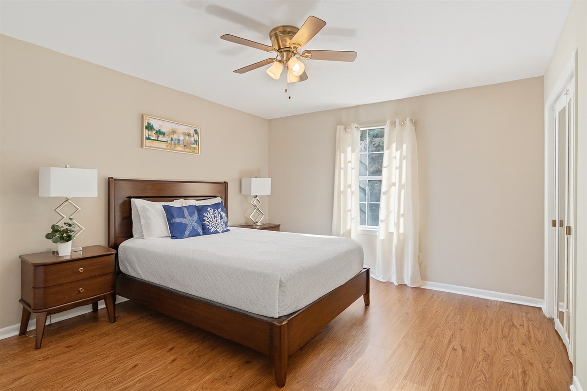 This bright and cozy bedroom features a queen bed with elegant coastal-themed accents, two matching nightstands, and a ceiling fan for added comfort. Natural light flows in through the window, creating a warm and inviting atmosphere perfect for rest and re