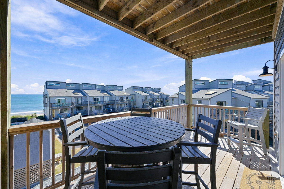 Covered wooden balcony setup with a high-top round dining table, matching chairs, an additional elevated captain's chair, and scenic ocean and coastal community views.