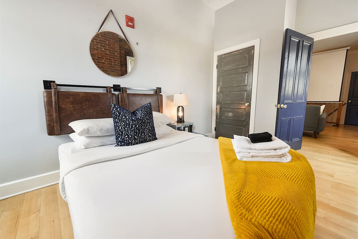 Relax in this bedroom featuring a white bed, small TV, and exposed brick walls for a modern touch. Wake up to natural light from the wide window, creating a bright and cozy ambiance. The bedside lamp sets the perfect mood for a peaceful night’s rest.