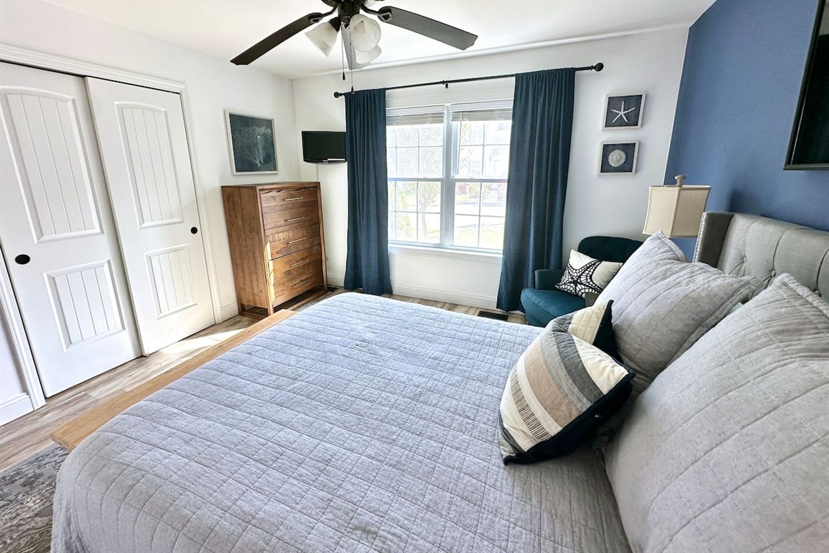 This bedroom offers a cozy layout with a large window. It features a tall wooden dresser, a double-door closet, and a comfortable bed. A blue accent wall and teal armchair add a stylish touch to the space.