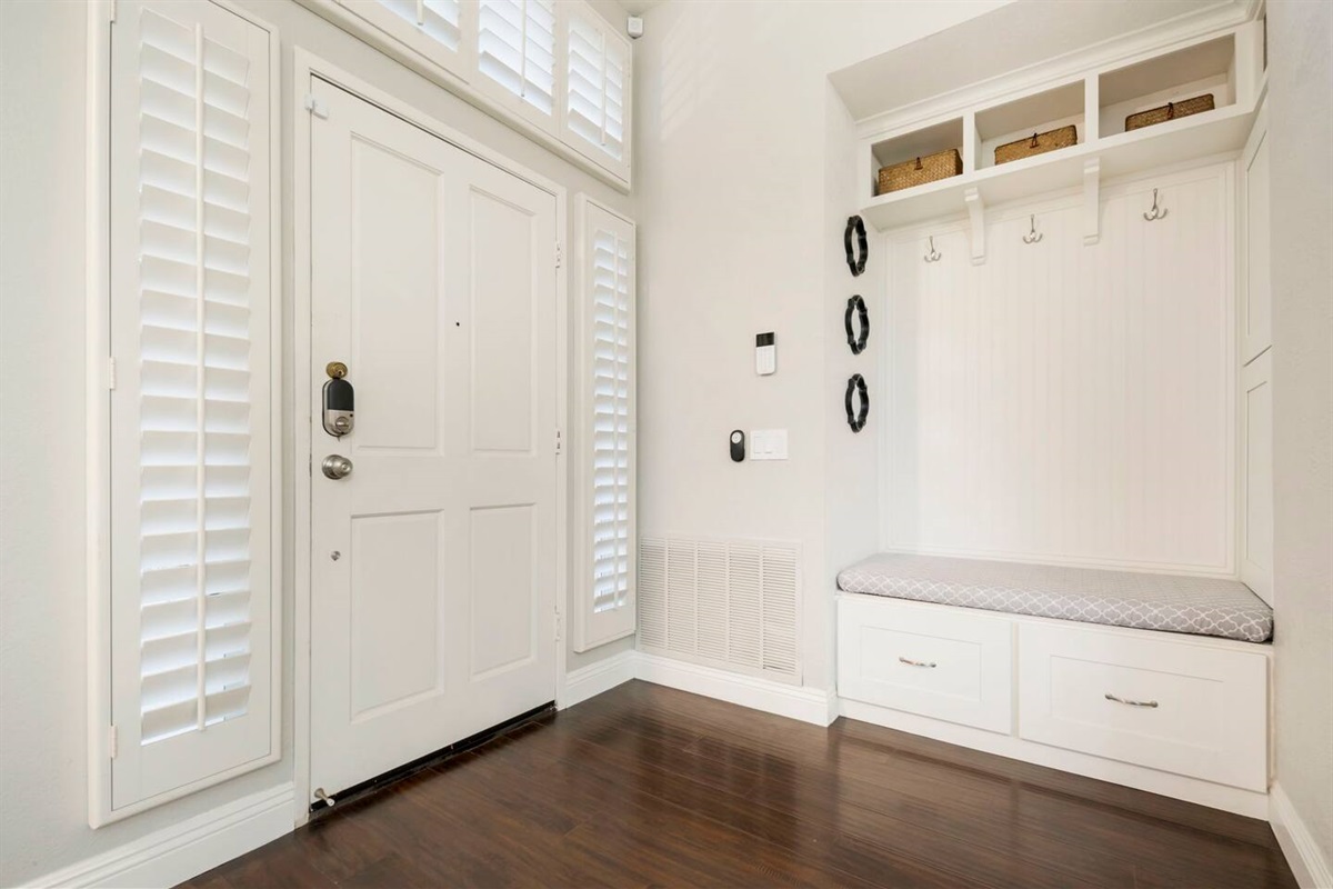 An inviting entrance door opens to a mudroom with ample storage, featuring built-in cabinets and shelves for shoes and bags. Hanging hooks line the walls for coats and hats, creating a tidy, organized space that’s both functional and welcoming.