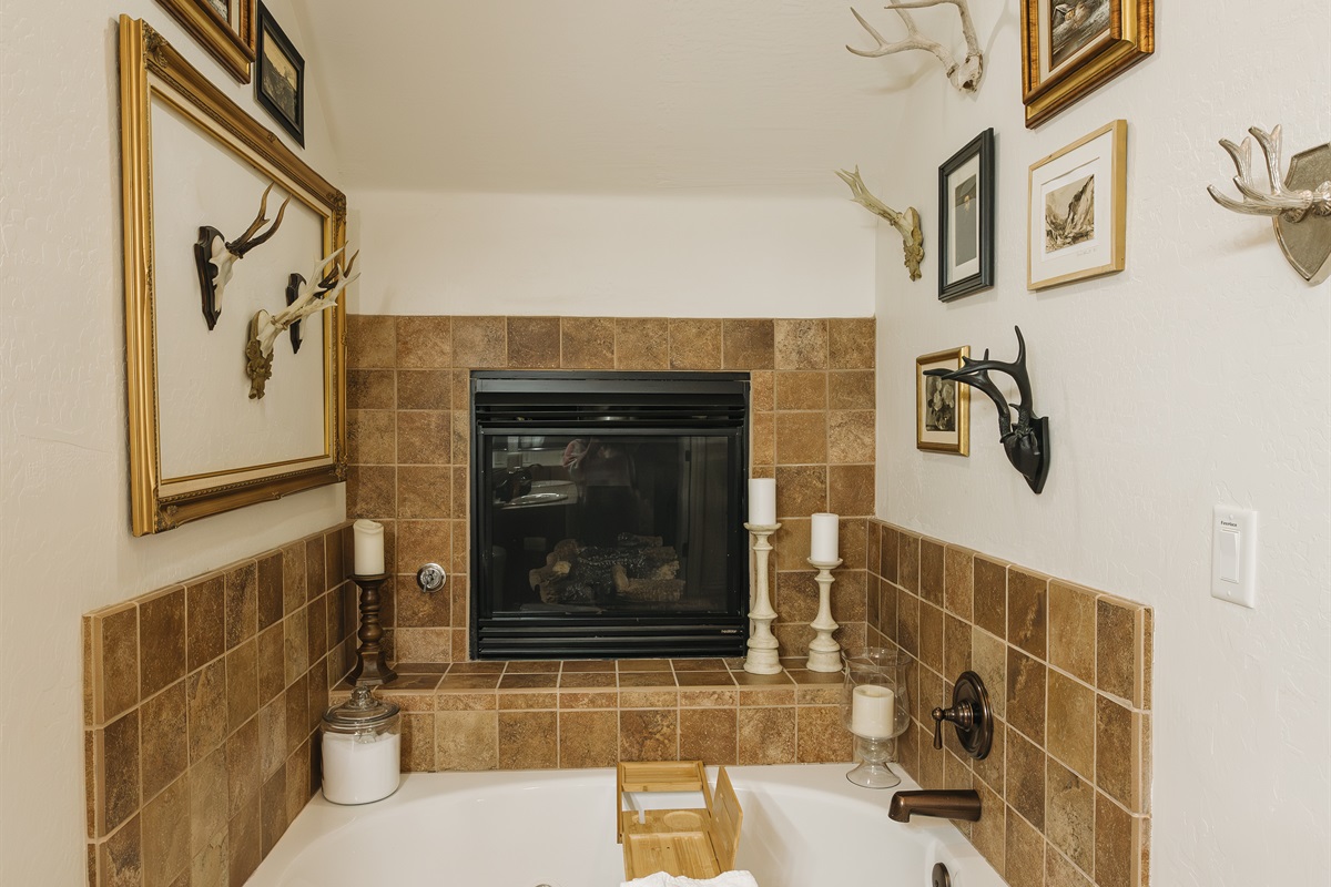 Soak beneath a glowing fireplace in this spa-like bathroom retreat. The deep jetted tub is framed by warm stone tile, curated vintage accents, plush towels, and soft lighting—creating a serene, romantic space to unwind after a day in the White Mountains.