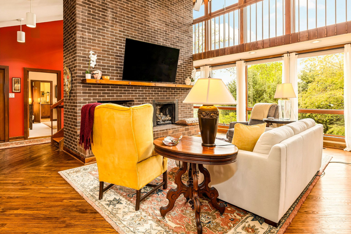 Light-filled Great Room featuring a two-story brick fireplace, handrailed catwalk, and large windows. Spacious comfortable seating with treetop views and primary Mountain View suite beyond.