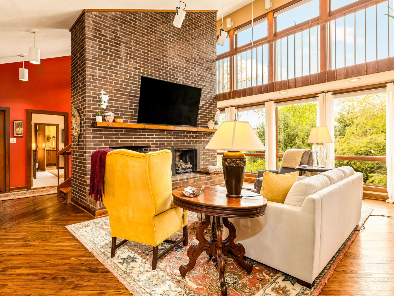 Light-filled Great Room featuring a two-story brick fireplace, handrailed catwalk, and large windows. Spacious comfortable seating with treetop views and primary Mountain View suite beyond.