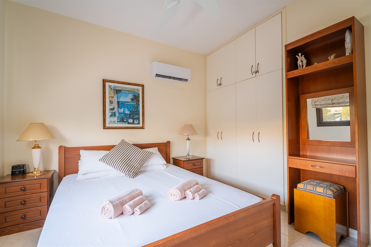 Double bedroom with a window facing the front of the villa. The room has spacious closets, a ceiling fan, a dedicated AC/heating unit, and a Bluetooth-enabled speaker/alarm clock combo.