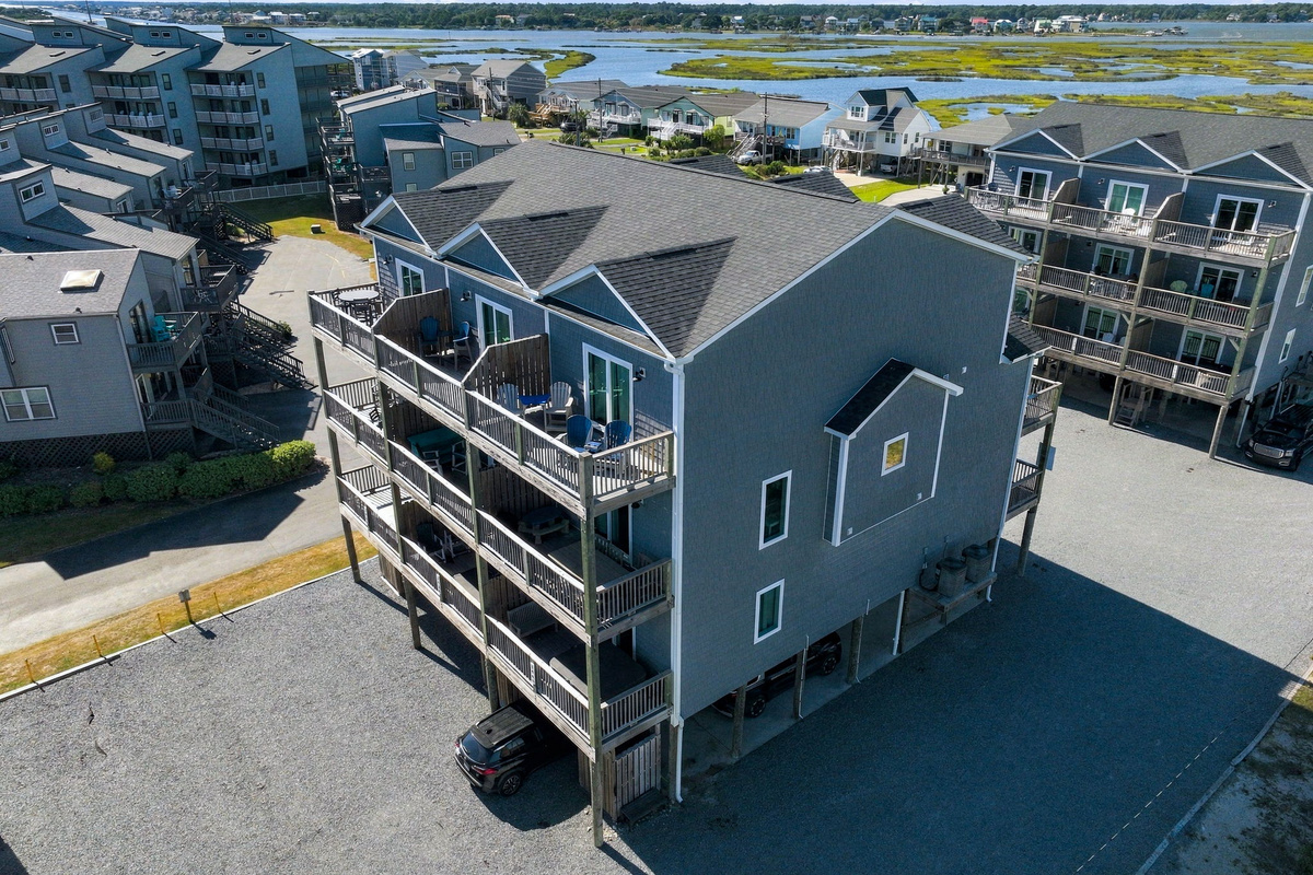 Wide-angle aerial view of the townhouse exterior showcasing the architectural design, multi-level private decks, covered ground-level parking, and surrounding coastal community scenery.   