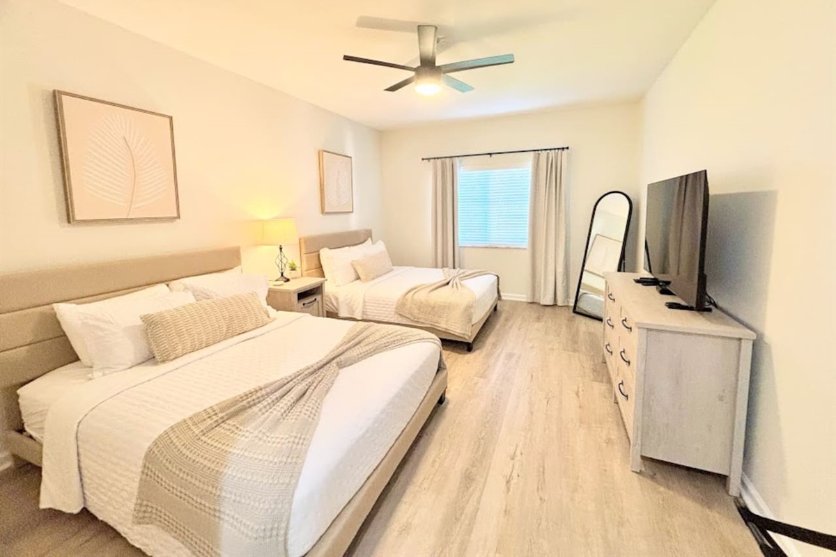 Bright and modern bedroom featuring two comfortable beds, wood-look flooring, ceiling fan, dresser storage, and a TV for a relaxing stay.