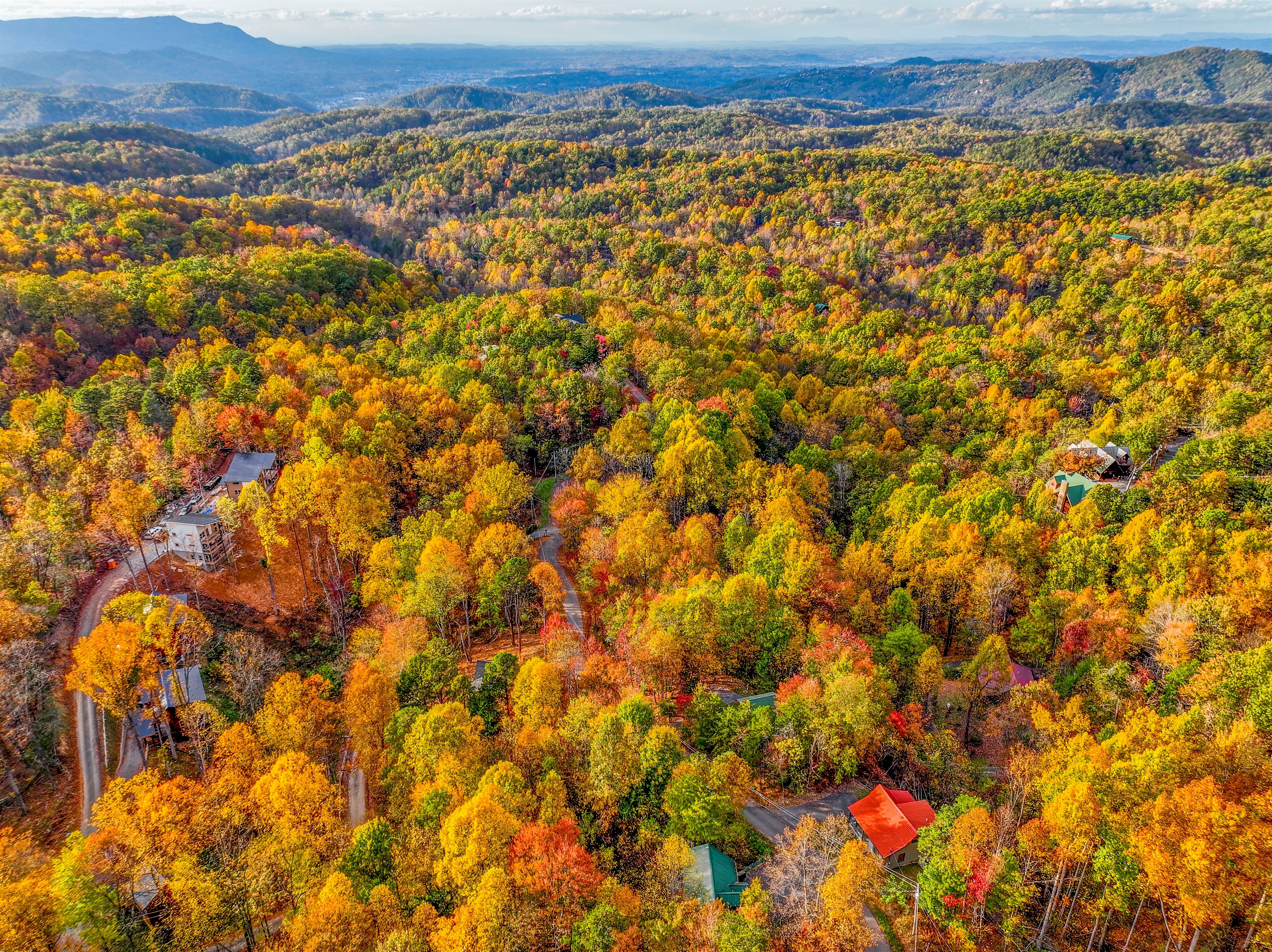 An aerial view of a serene autumn landscape, where vibrant hues of gold, orange, and red blanket the forest. Cozy cabins nestle among the trees, with majestic mountains rising in the distance.