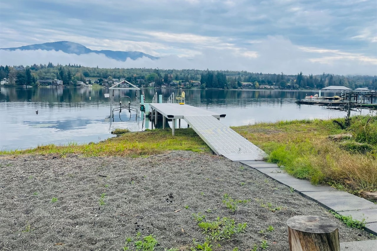 A dock stretches into the calm lake, accessible by a pathway with sand on one side and grass on the other. It’s perfect for mooring a boat or sitting by the water, blending natural textures with the peaceful lakeside setting.