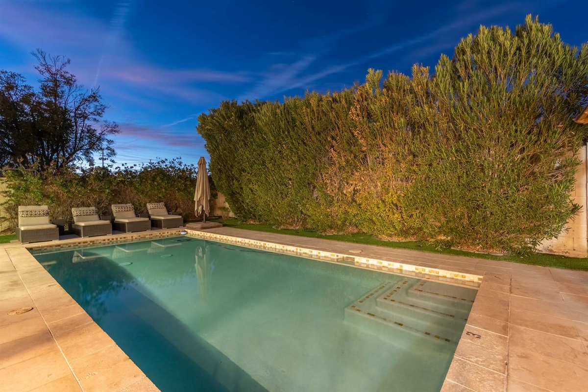 Relax in style by the serene pool, beautifully lit under the twilight sky, with privacy hedges and comfortable loungers creating a peaceful retreat.