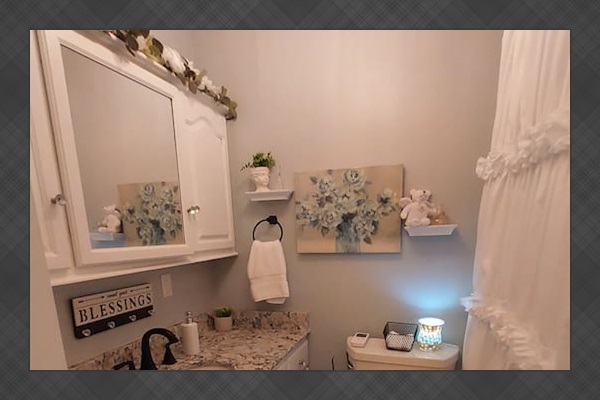 Pale blue walls offer a refined contrast to the crisp white décor, with a romantic ruffled shower curtain and vintage-style vanity. A sparkling marbled countertop adds beauty and space, creating a serene, elegantly styled sanctuary.