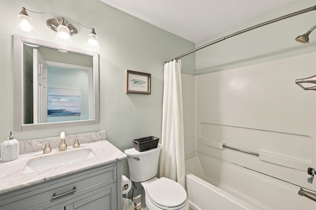 Primary bathroom suite featuring a single vanity with gray cabinetry, marble-look countertop, undermount sink, three-light sconce vanity fixture, framing mirror, soft seafoam walls, and full shower-tub combination.   
