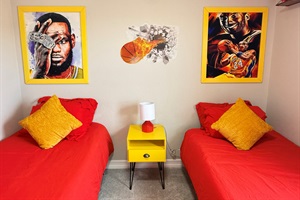Closer look at the upstairs twin bedroom with two twin beds, a shared nightstand with a lamp between them, and sports themed artwork above.