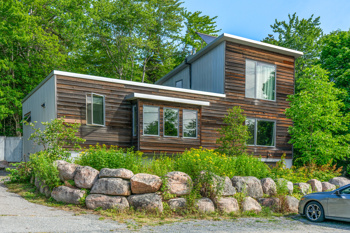 The Crag" emerges from natural granite boulders on the Acadia border. This three-level architectural sanctuary features rich wood siding, corrugated metal accents, and massive windows framed by lush forest. Steps to Cadillac Mountain trails.