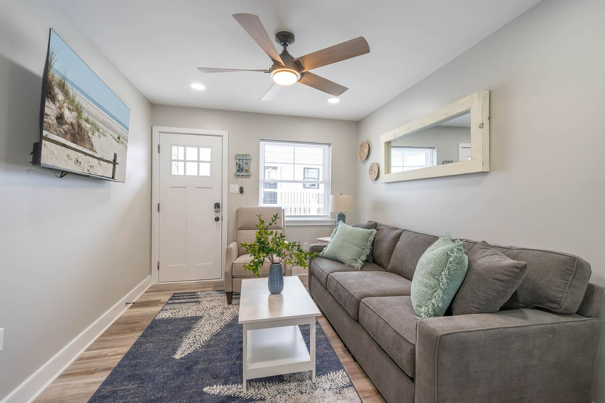 Bright and comfortable living room with plush seating, ceiling fan, and natural light from the front window. Soft coastal colors and a smart TV create a relaxed space to unwind, read, or enjoy a quiet evening indoors.