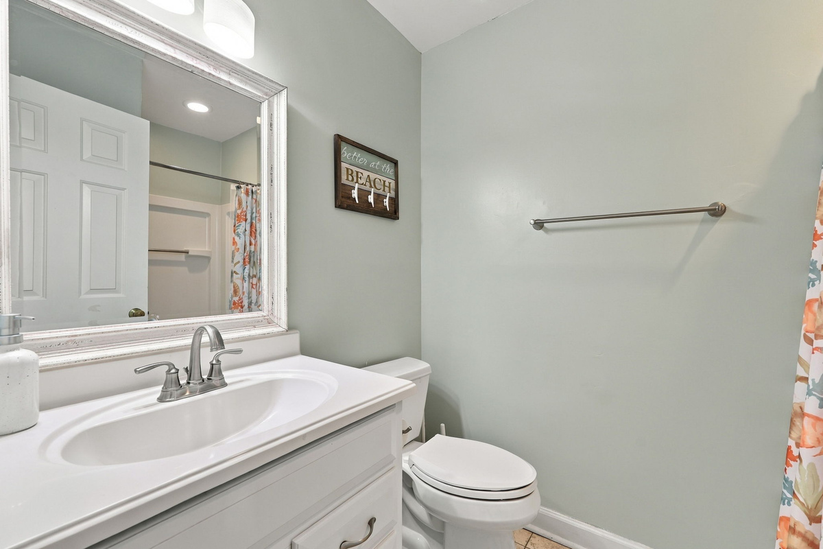 Refined secondary bathroom featuring a crisp white vanity with drop-in sink, brushed nickel fixtures, a distressed white-framed mirror, soft seafoam walls, and playful coastal wall accents.   