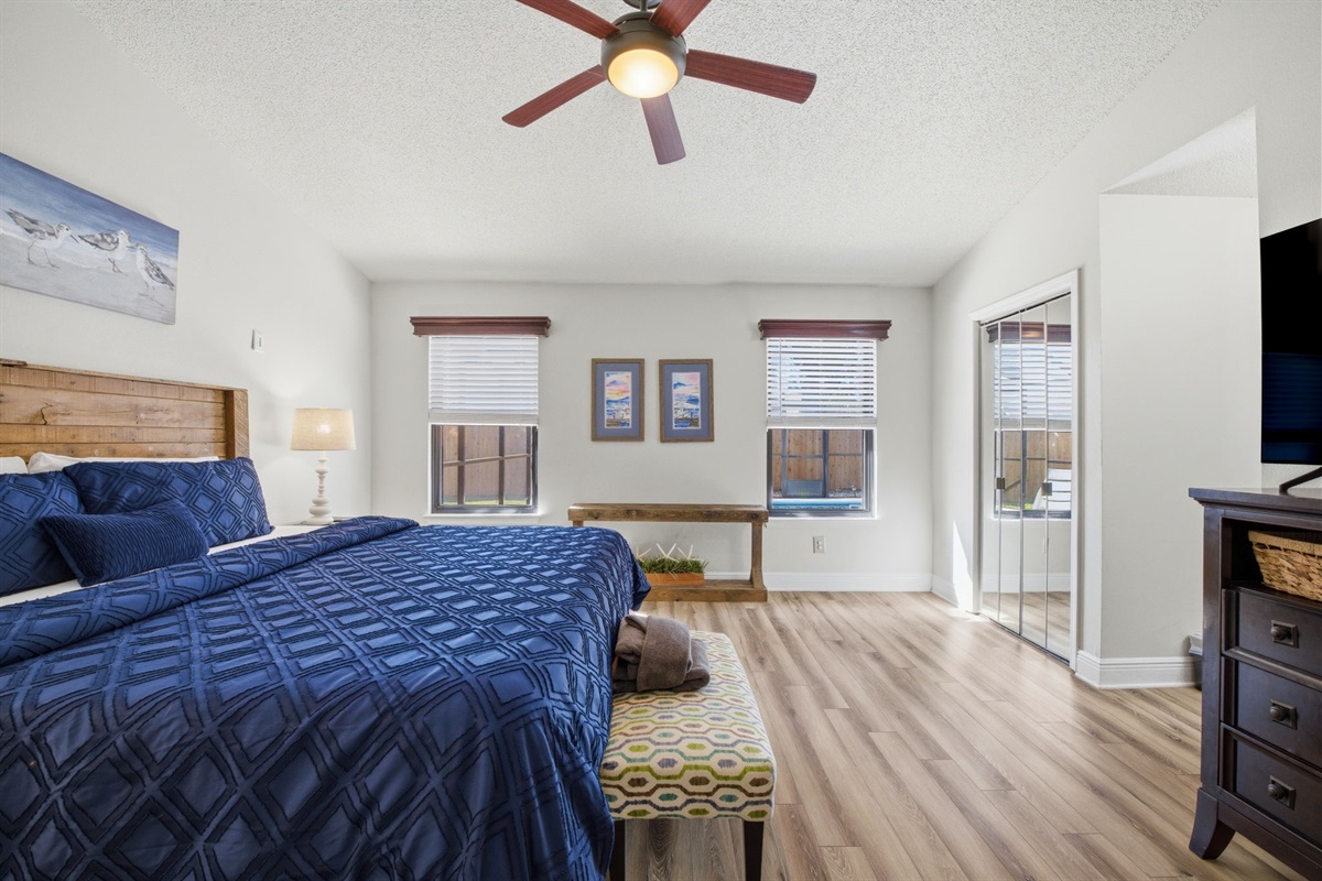 This spacious bedroom offers a relaxing retreat with a comfortable king-size bed, soft lighting, and plenty of natural light from the surrounding windows. The open layout and calming coastal décor create a bright and peaceful place to unwind.