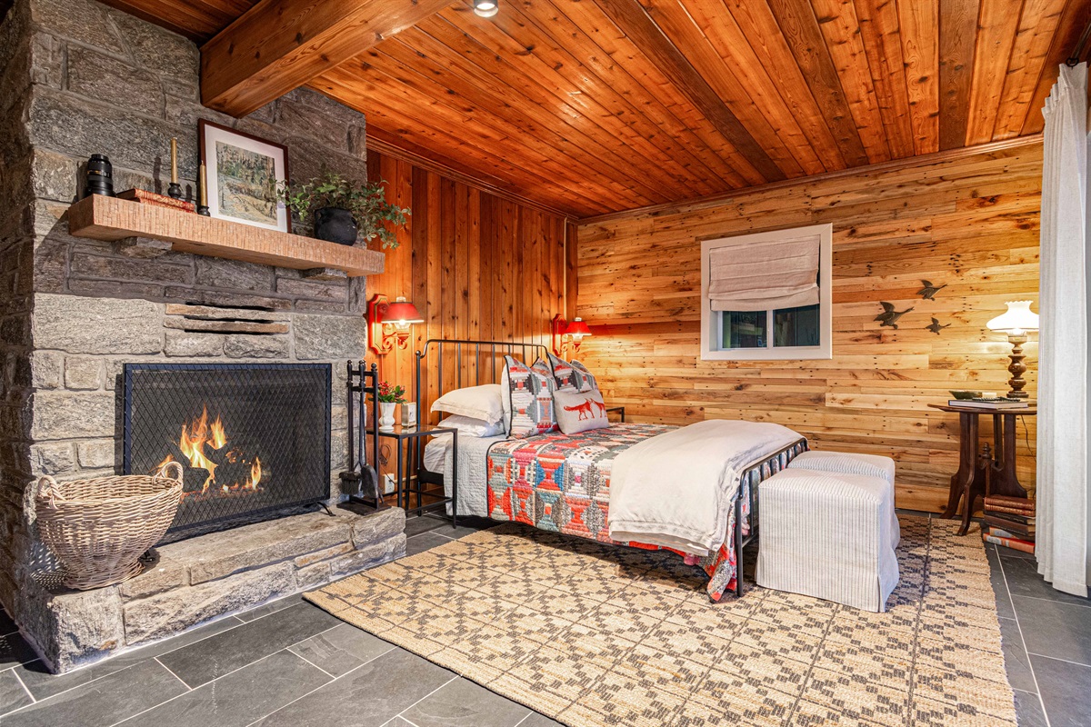 A cozy primary bedroom retreat where rustic charm meets comfort, featuring a stone fireplace, warm wood walls, and a plush bed designed for restful mountain nights.
