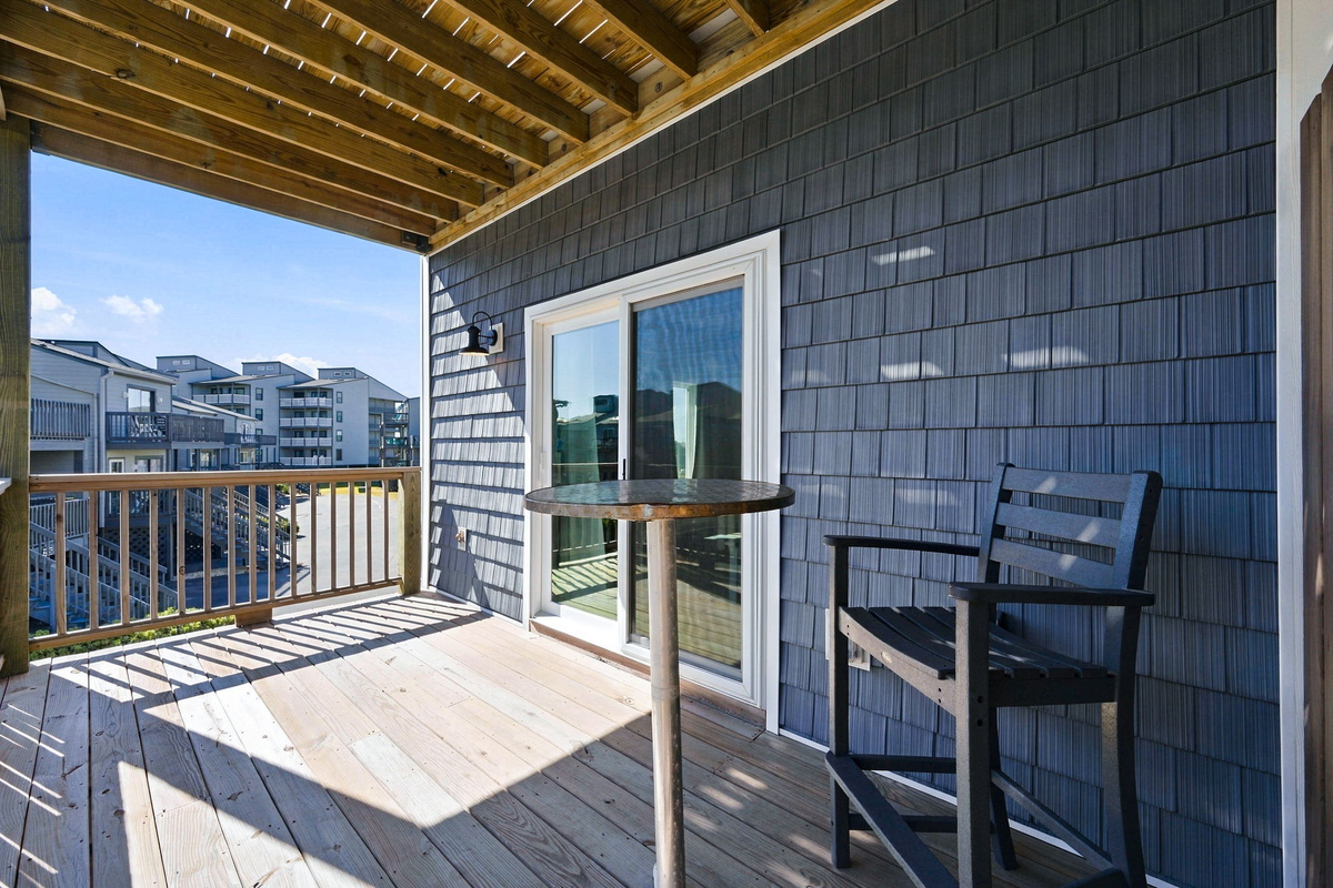 Covered wooden balcony furnished with a high-top bistro table and chair, offering a shaded outdoor seating area accessible via sliding glass doors.   