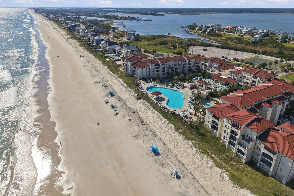 Aerial coastline view detailing the sandy beach, surf line, resort pool courtyard, and surrounding coastal community set between the ocean and sound.