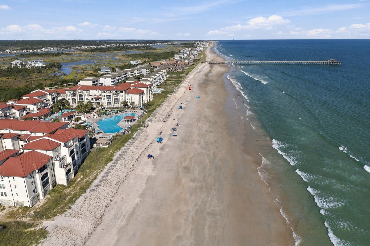 Elevated shoreline view detailing the beachfront resort grounds, wide sandy beach, surf line, and long pier extending into the ocean.   