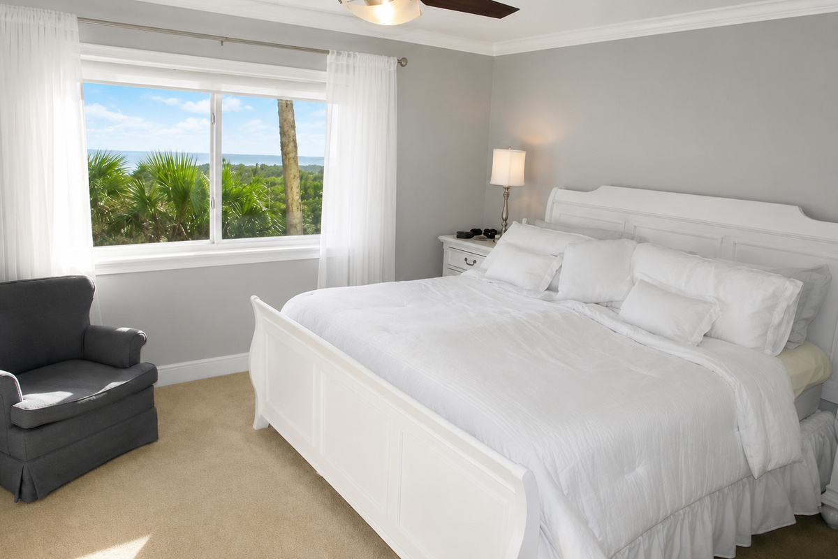 Bright bedroom with white bedding, gray armchair, and serene palm tree view outside.