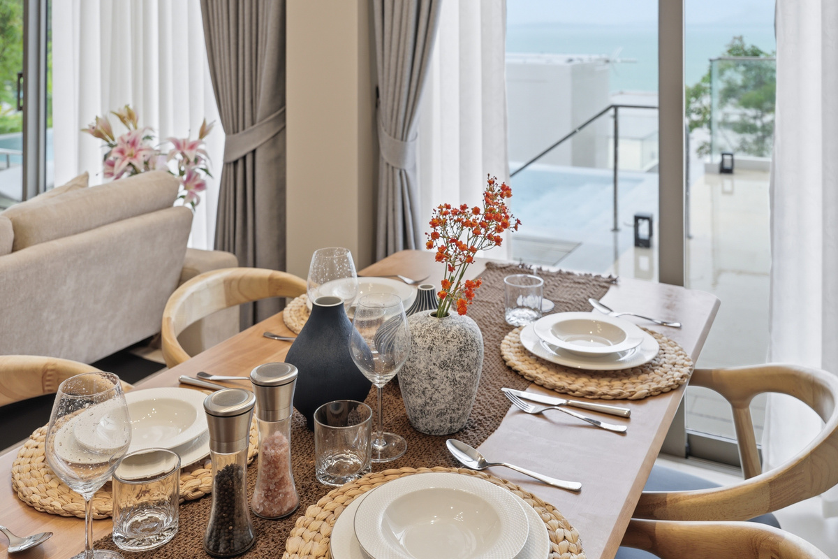 Dining area with ocean view, wooden table set for two, beige curtains, and coastal decor.