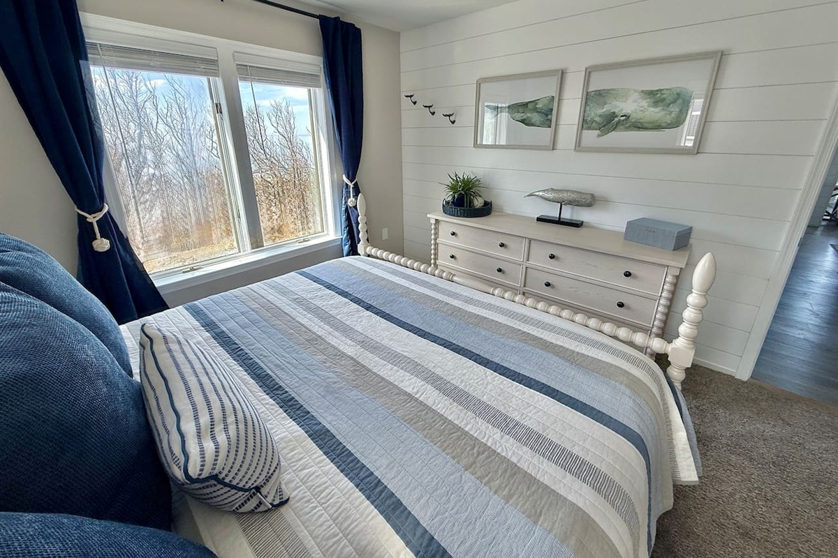 Enjoy the coastal-themed bedroom features a bed with blue-striped bedding, deep blue curtains, a light-toned dresser, and nautical wall art, creating a tranquil and well-coordinated seaside retreat.