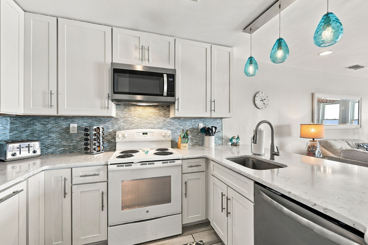 Bright coastal kitchen feature showcasing vibrant aqua pendant lighting, shimmering blue mosaic tile backsplash, classic white shaker cabinetry, polished white quartz countertops, an undermount sink with a high-arc faucet, and an electric range setup with 